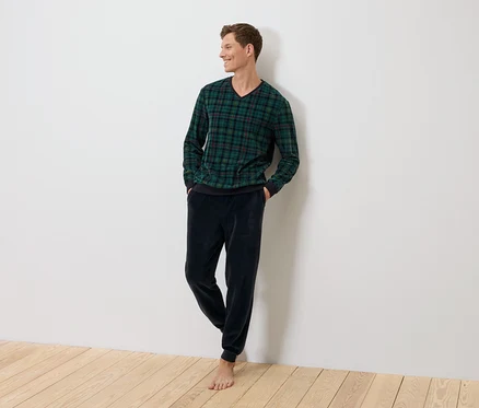 Ein Mann steht an einer Wand, bekleidet mit einem grün karierten Pullover und dunkelblauen Samthosen.