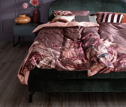 Grünes Bett mit Blumendesign-Bettwäsche und Kissen. Ein Nachttisch mit Vase und Lampe steht neben dem Bett.