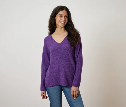 Eine lächelnde Frau mit langen, gewellten Haaren trägt einen lilafarbenen Pullover mit V-Ausschnitt und Jeans.