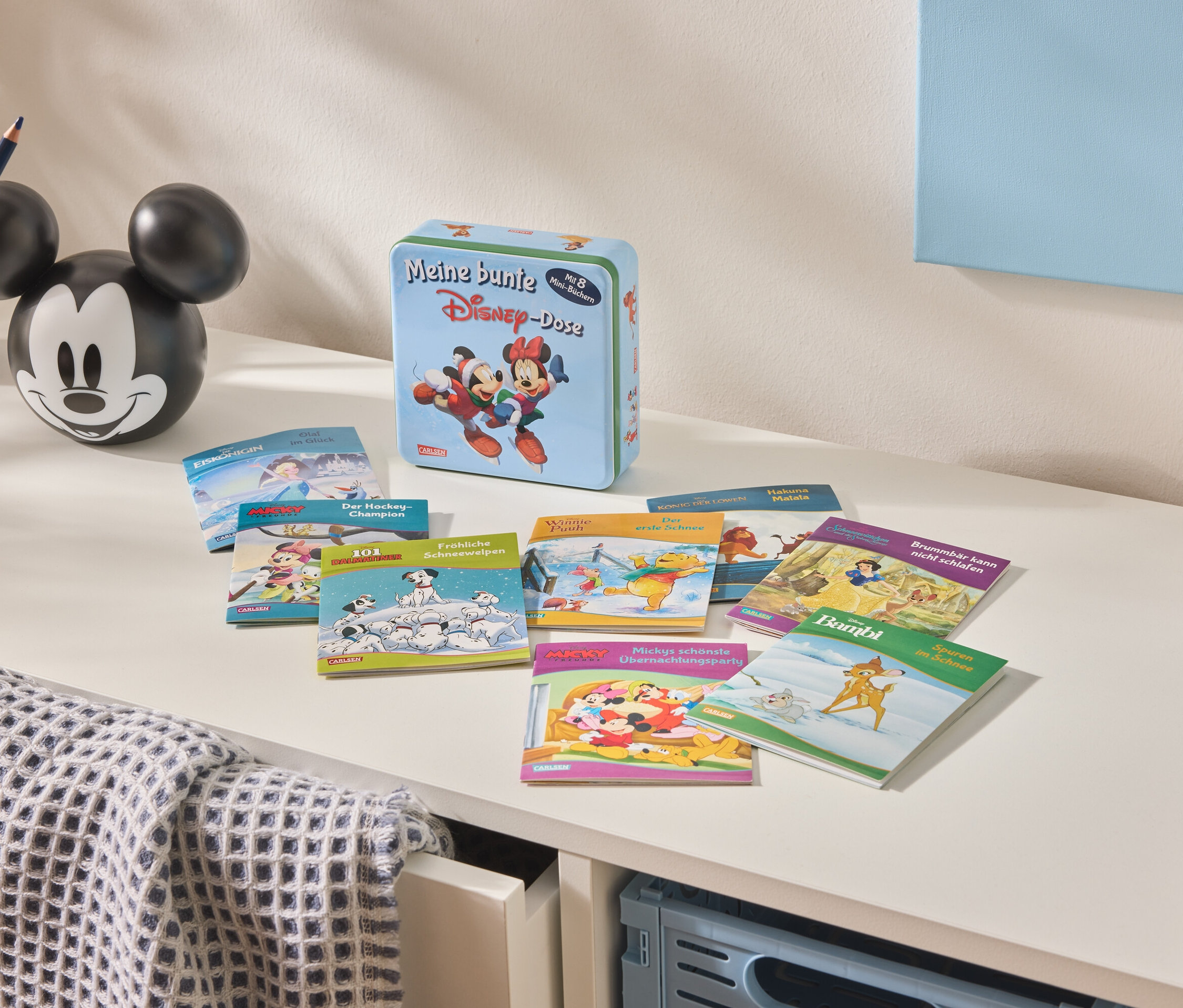 Auf einer weißen Kommode liegen eine Disney-Blechdose, Kinderbücher und eine Vase mit Buntstiften.