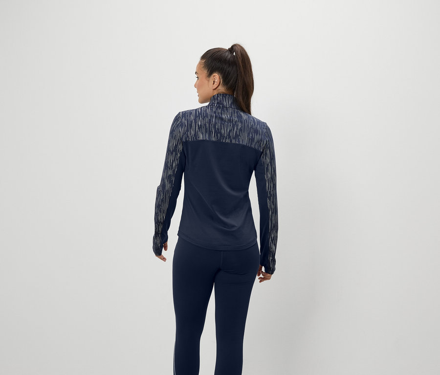 Frau mit braunen Haaren, die zu einem Pferdeschwanz zusammengebunden sind, steht mit dem Rücken zur Kamera und trägt eine dunkelblaue Jacke und Hose.