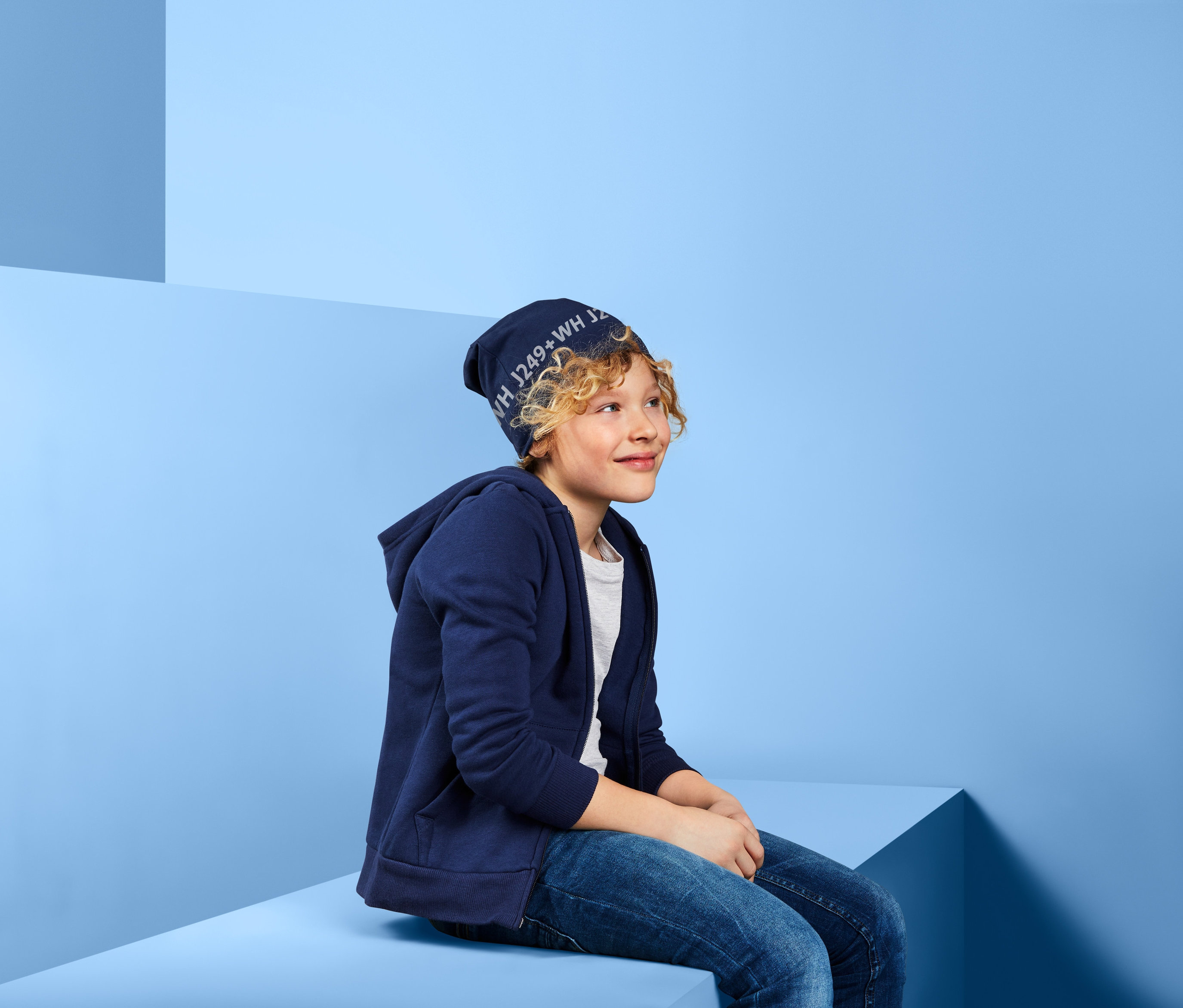 Ein Junge mit blonden Haaren sitzt und trägt eine Wendemütze und eine blaue Kapuzenjacke.