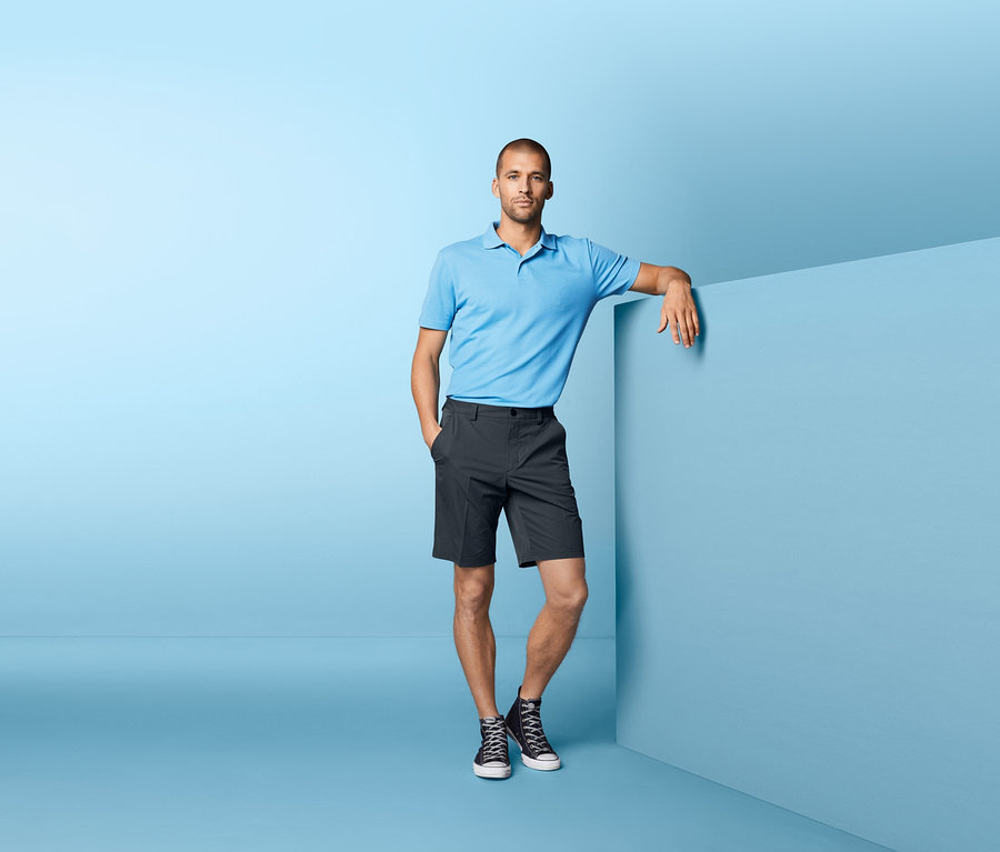 Mann in blauem Poloshirt und Funktionsshorts lehnt an einer blauen Wand.