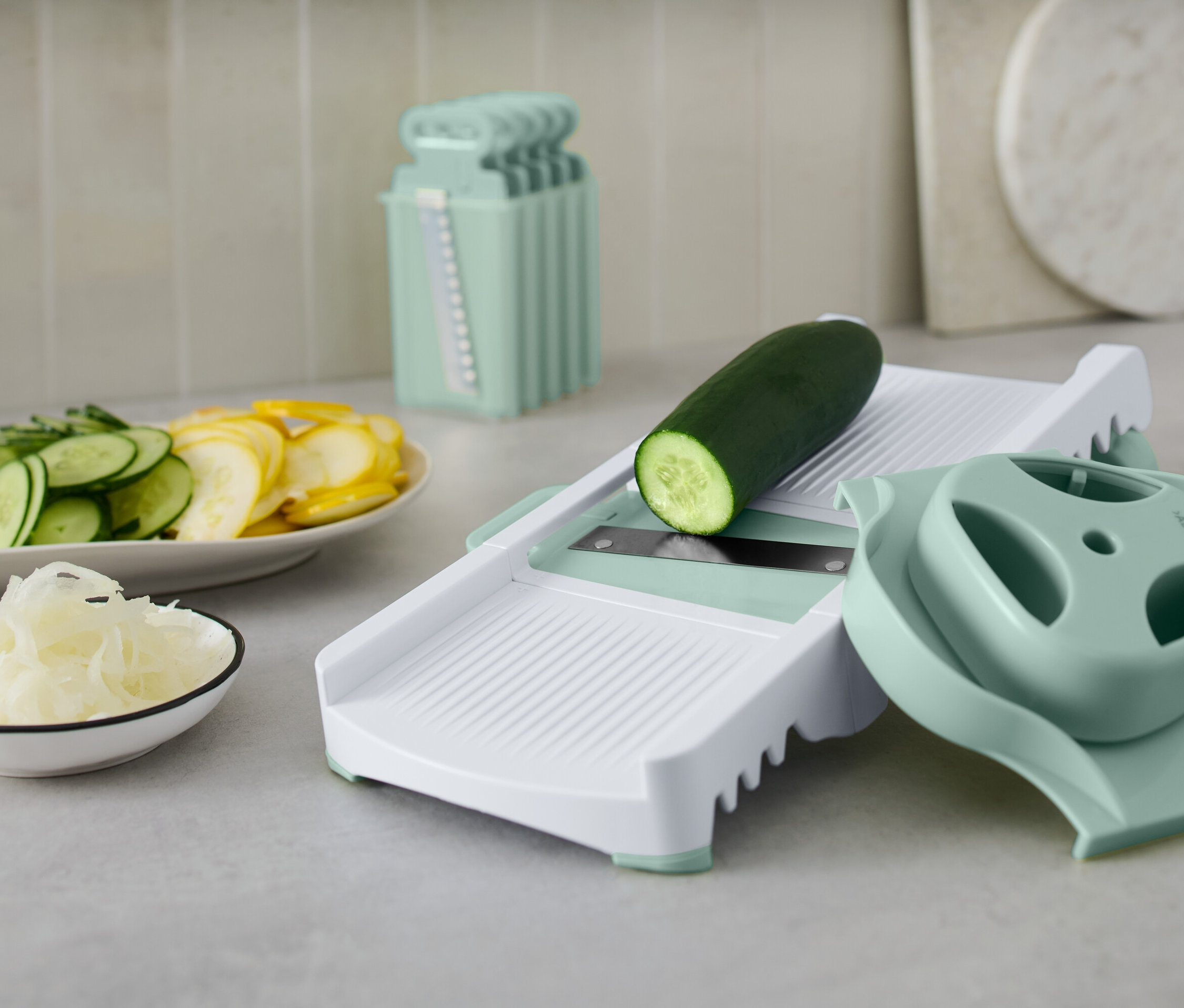 Eine grüne Gurke auf einem weißen Gemüseschneider mit Zubehör. Auf einem Teller liegen geschnittene Gurken und gelber Kürbis.