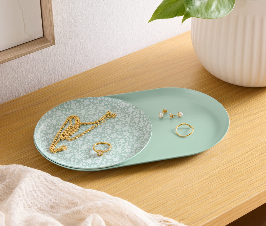 Auf einem Holzregal liegt eine grüne Schale mit einer goldenen Halskette, einem Ring und Ohrringen. Daneben steht eine weiße Vase.