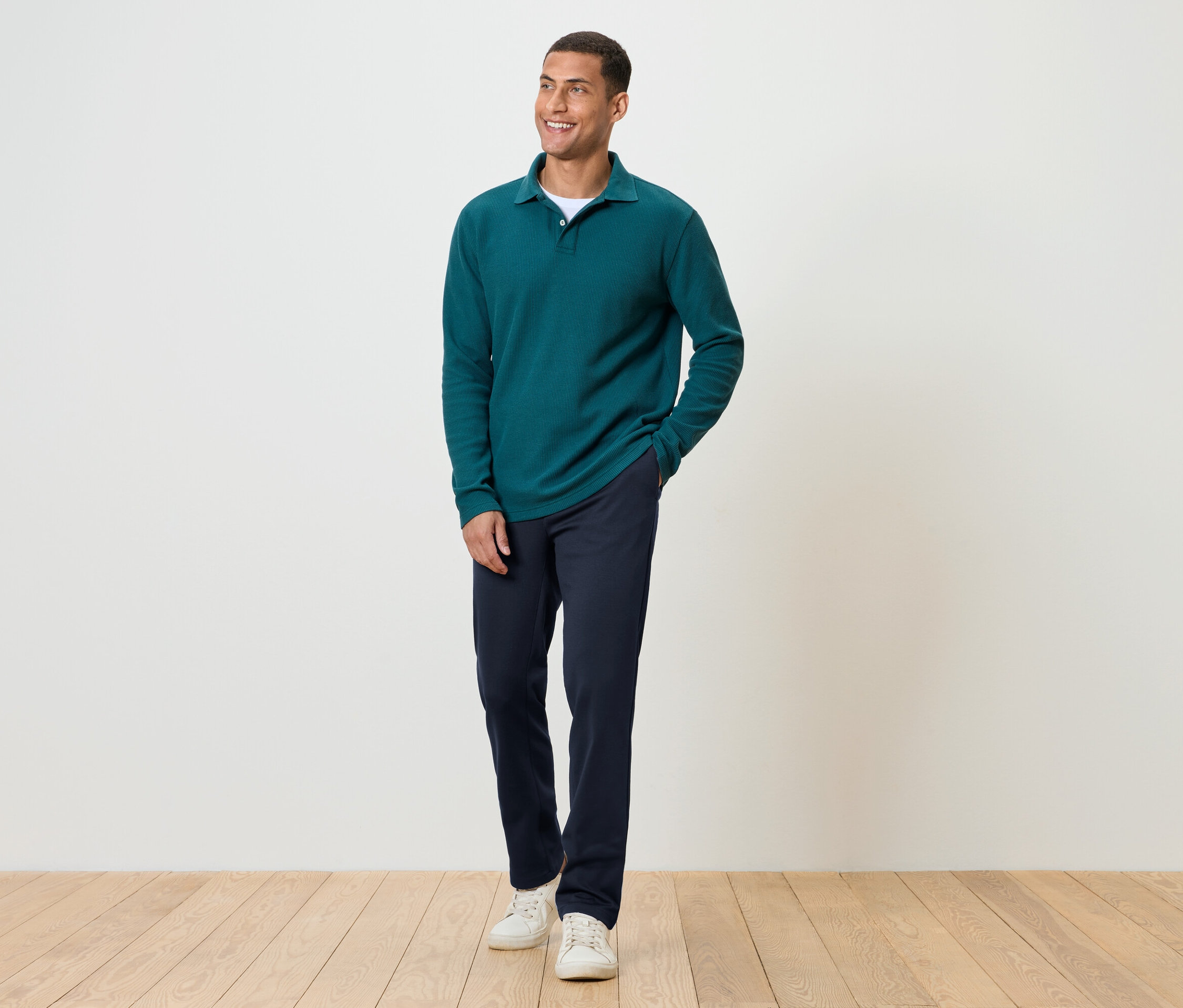 Ein Mann steht auf einem Holzboden und trägt eine dunkelblaue Hose, ein grünes Langarmshirt und weiße Turnschuhe.