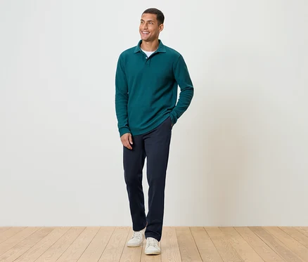Ein Mann steht auf einem Holzboden und trägt eine dunkelblaue Hose, ein grünes Langarmshirt und weiße Turnschuhe.