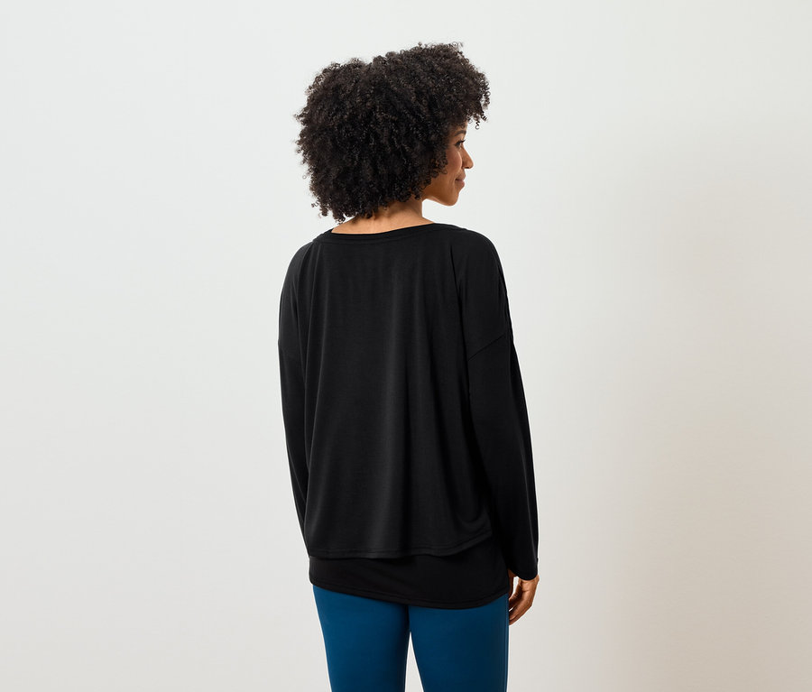 Von hinten ist eine Frau mit lockigem Haar zu sehen, die ein schwarzes Langarmshirt und blaue Leggings trägt.