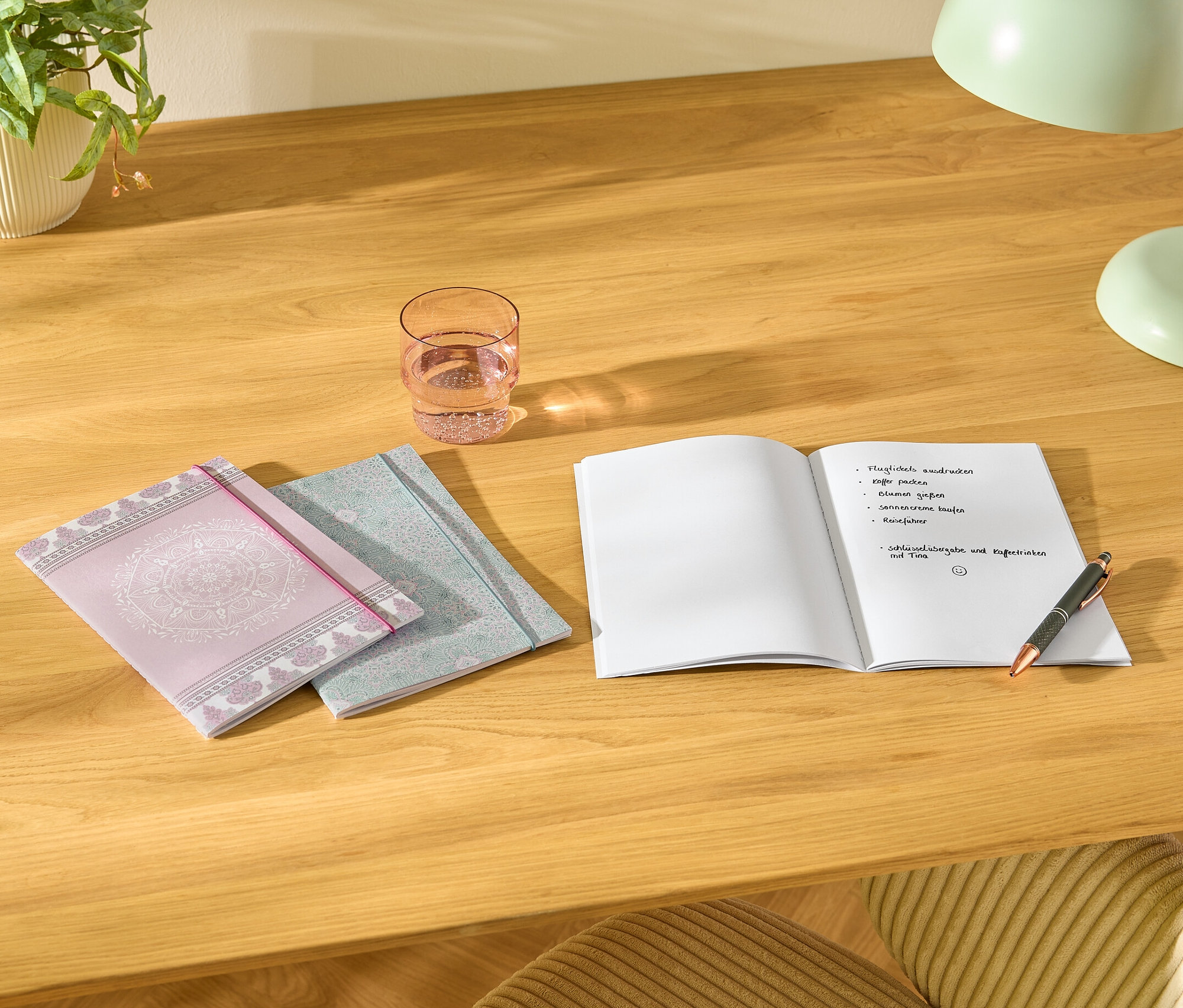 Auf einem Tisch liegen drei Notizbücher, ein Glas Wasser, ein offenes Notizbuch mit Notizen und ein Stift.