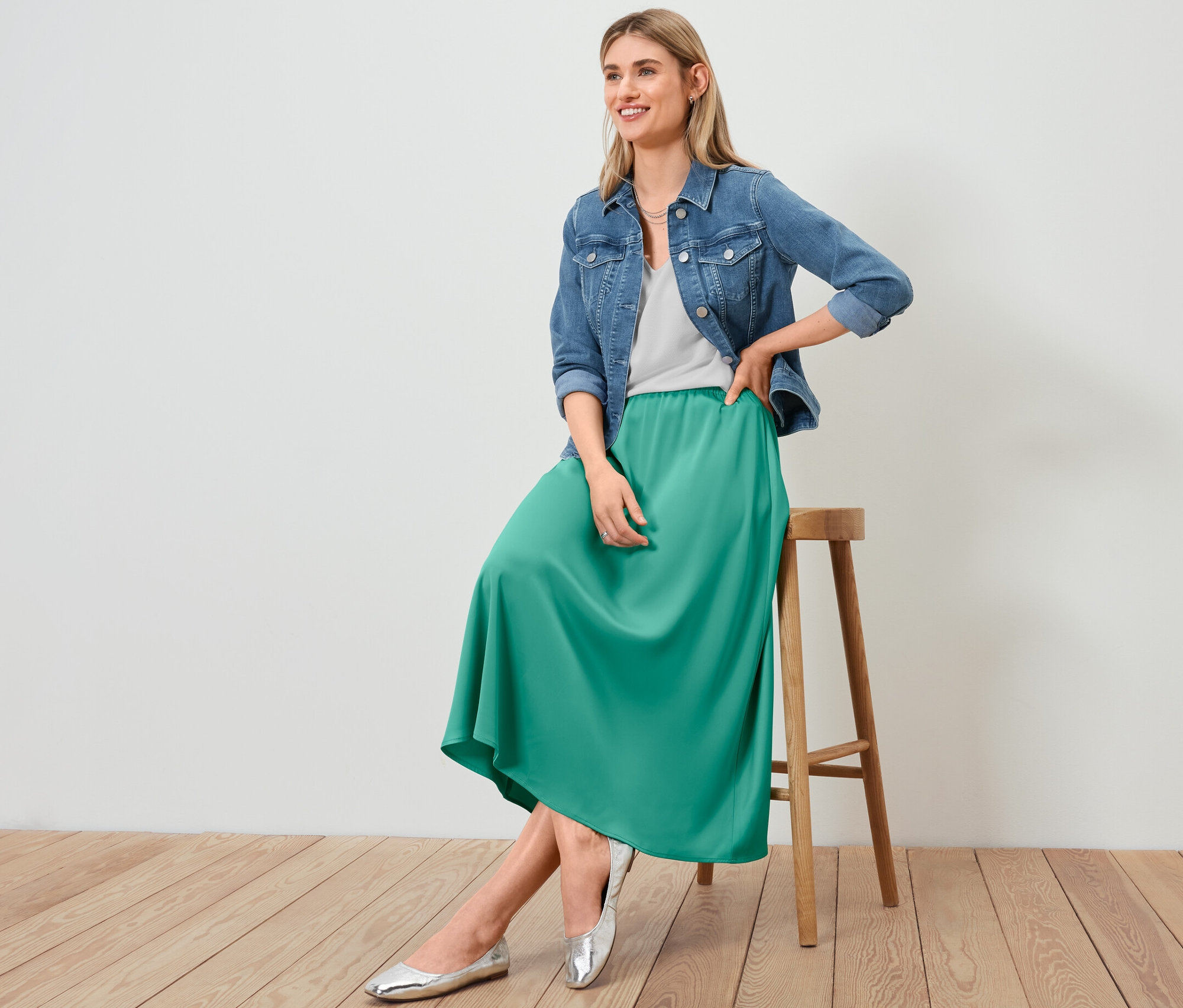 Frau sitzt auf einem Hocker und trägt eine Jeansjacke, einen Satinrock und ein strukturiertes Shirt in Weiß.