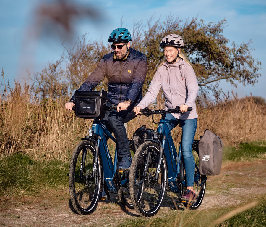 Ein Mann und eine Frau fahren mit ihren blauen E-Bikes durch die Natur. Beide tragen FISCHER Fahrradhelme Urban PLUS Chicago.