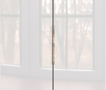 Nahaufnahme eines geschlossenen, weißen Schranks mit einem goldenen Griff vor einem Fenster.