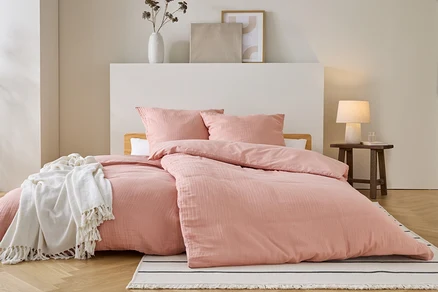 Ein Bett mit rosa Bettwäsche und Kissen steht auf einem gestreiften Teppich. Eine weiße Decke liegt auf dem Bett. Ein Nachttisch steht daneben.