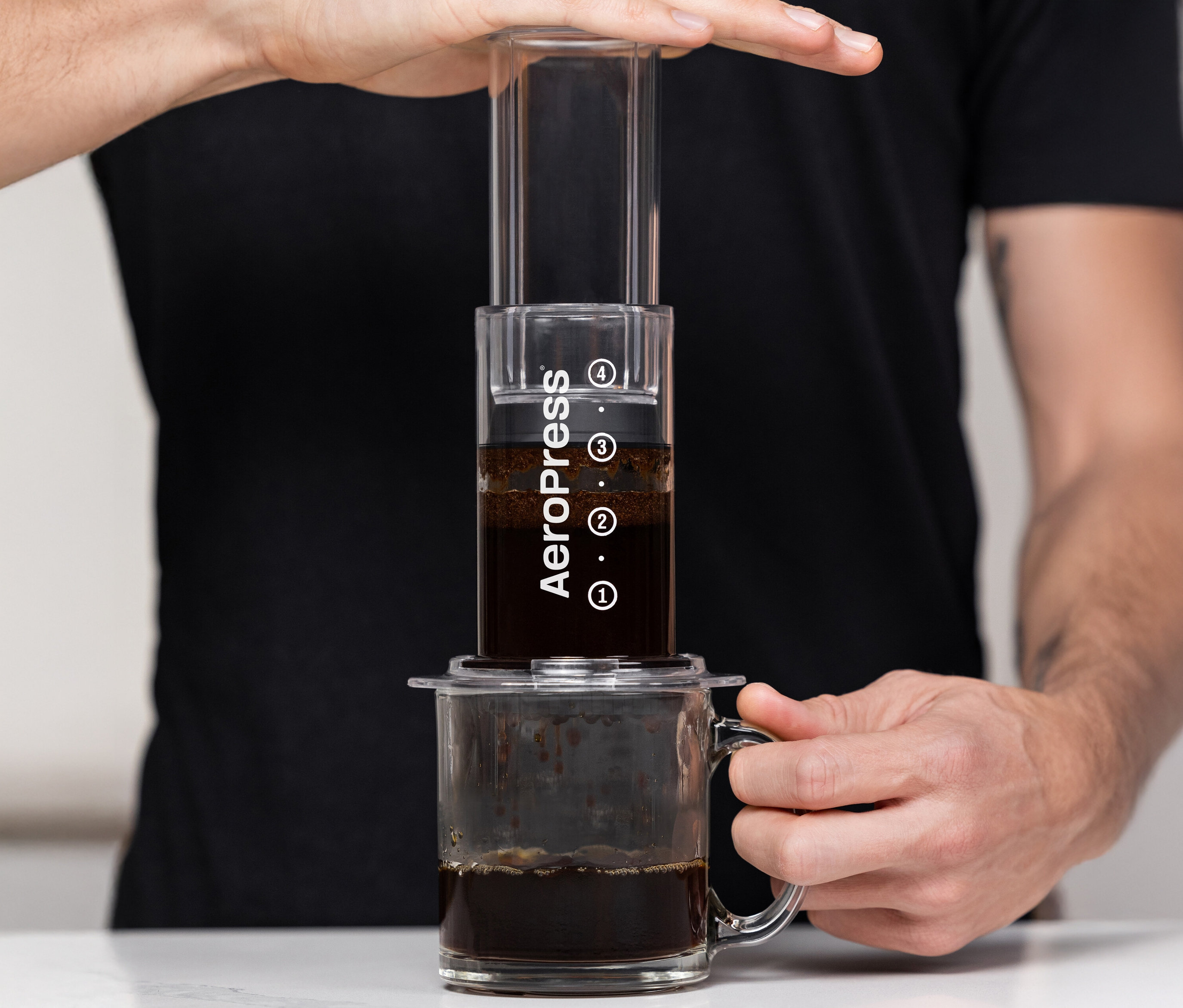 Zwei Hände bedienen eine durchsichtige AeroPress Kaffeemaschine »Clear« über einer Tasse.