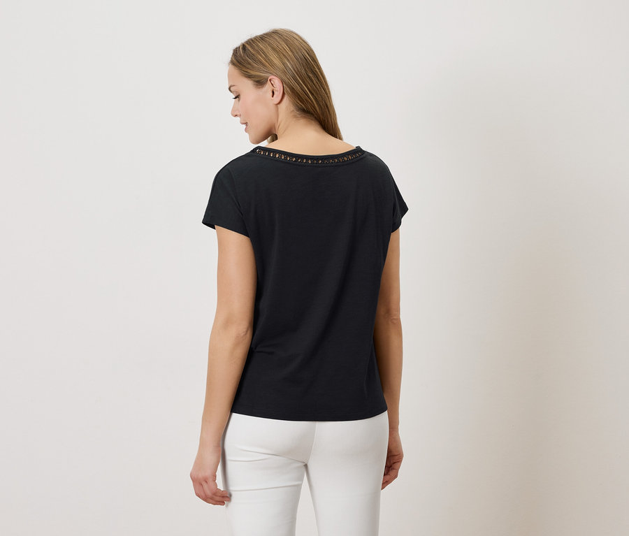 Frau von hinten, trägt ein schwarzes T-Shirt mit Lochmuster am Halsausschnitt und eine weiße Hose vor einem weißen Hintergrund.
