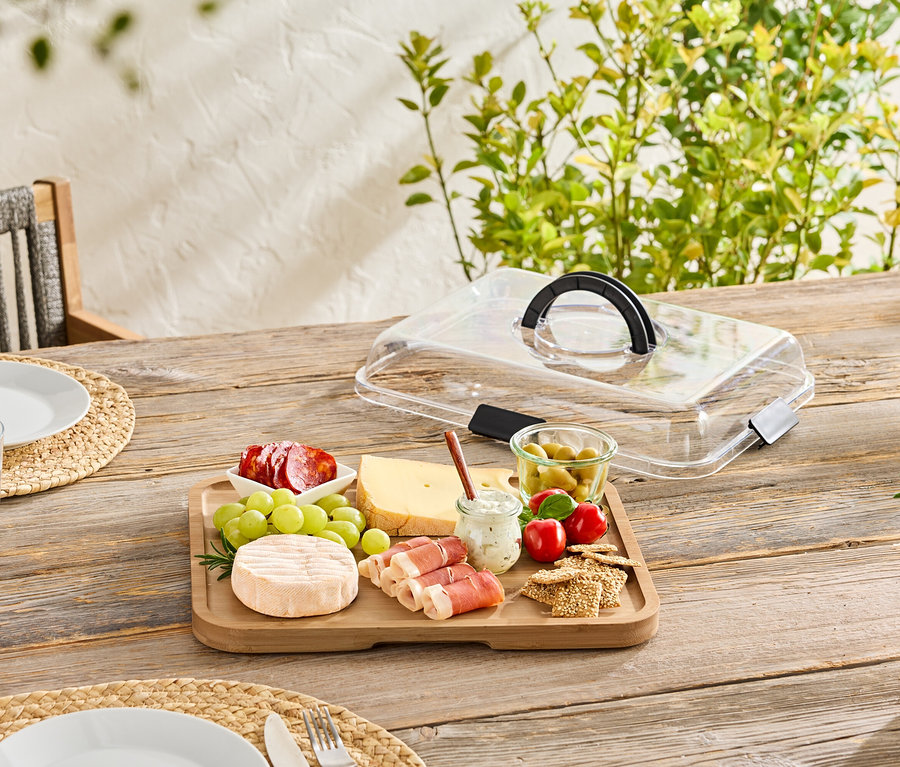 Auf einem Holztisch steht ein Holzbrett mit Käse, Wurst, Oliven und Cracker, teilweise mit einer transparenten Abdeckung bedeckt.