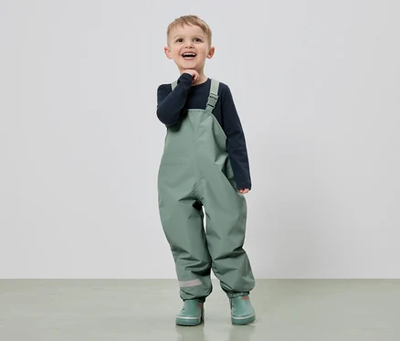 Ein lächelnder Junge in grüner Latzhose und Gummistiefeln steht vor einem hellen Hintergrund.