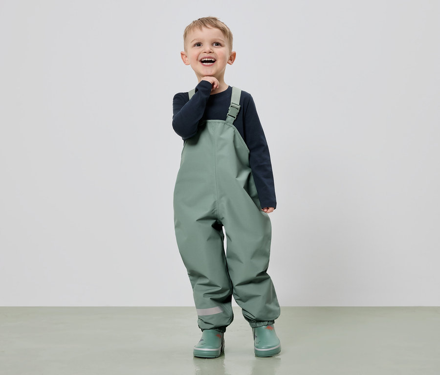 Ein lächelnder Junge in grüner Latzhose und Gummistiefeln steht vor einem hellen Hintergrund.
