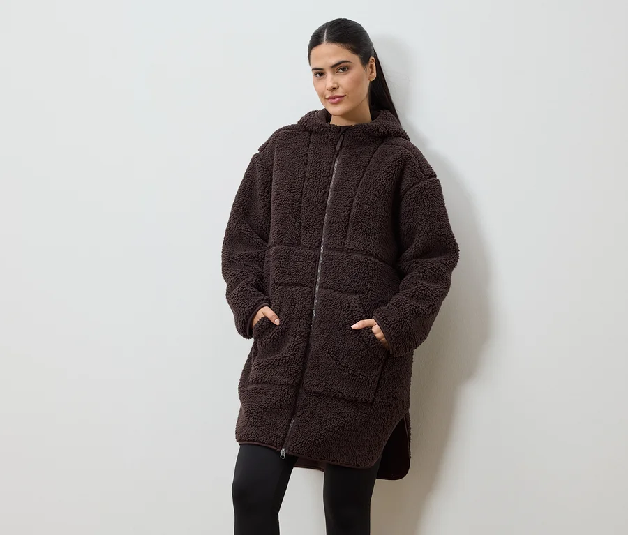 Frau mit Händen in den Taschen trägt einen langen, braunen Teddyfellmantel mit Kapuze und Reißverschluss über schwarzer Leggings.