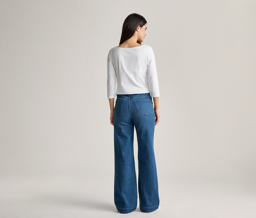 Rückansicht einer Frau mit dunklen Haaren, die ein weißes, langärmeliges Oberteil und eine weite, blaue Jeans trägt.
