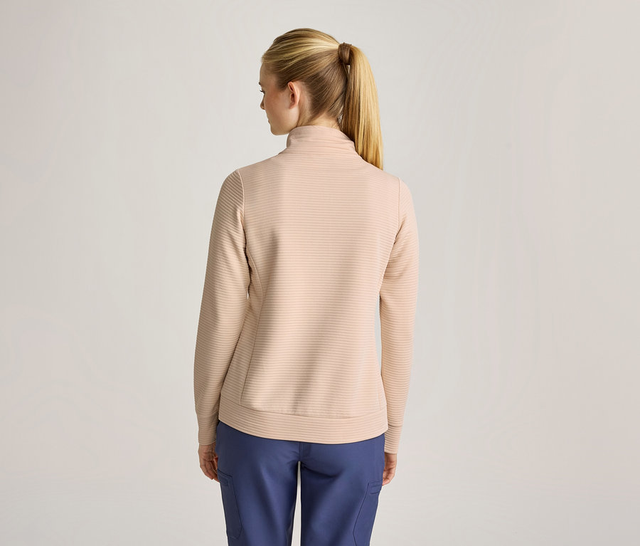 Eine Frau von hinten, mit blondem Pferdeschwanz, trägt ein beiges, geripptes Langarm-Oberteil mit Stehkragen und blaue Hosen.