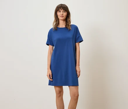 Frau trägt blaues T-Shirt-Kleid vor einem neutralen Hintergrund.