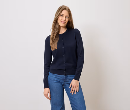 Frau mit langen braunen Haaren, trägt eine marineblaue Strickjacke mit Knöpfen und blaue Jeans vor einem hellen Hintergrund.