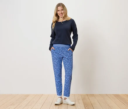 Eine blonde Frau steht vor einer weißen Wand, sie trägt einen dunkelblauen Pullover und blaue Hosen mit einem abstrakten Muster.