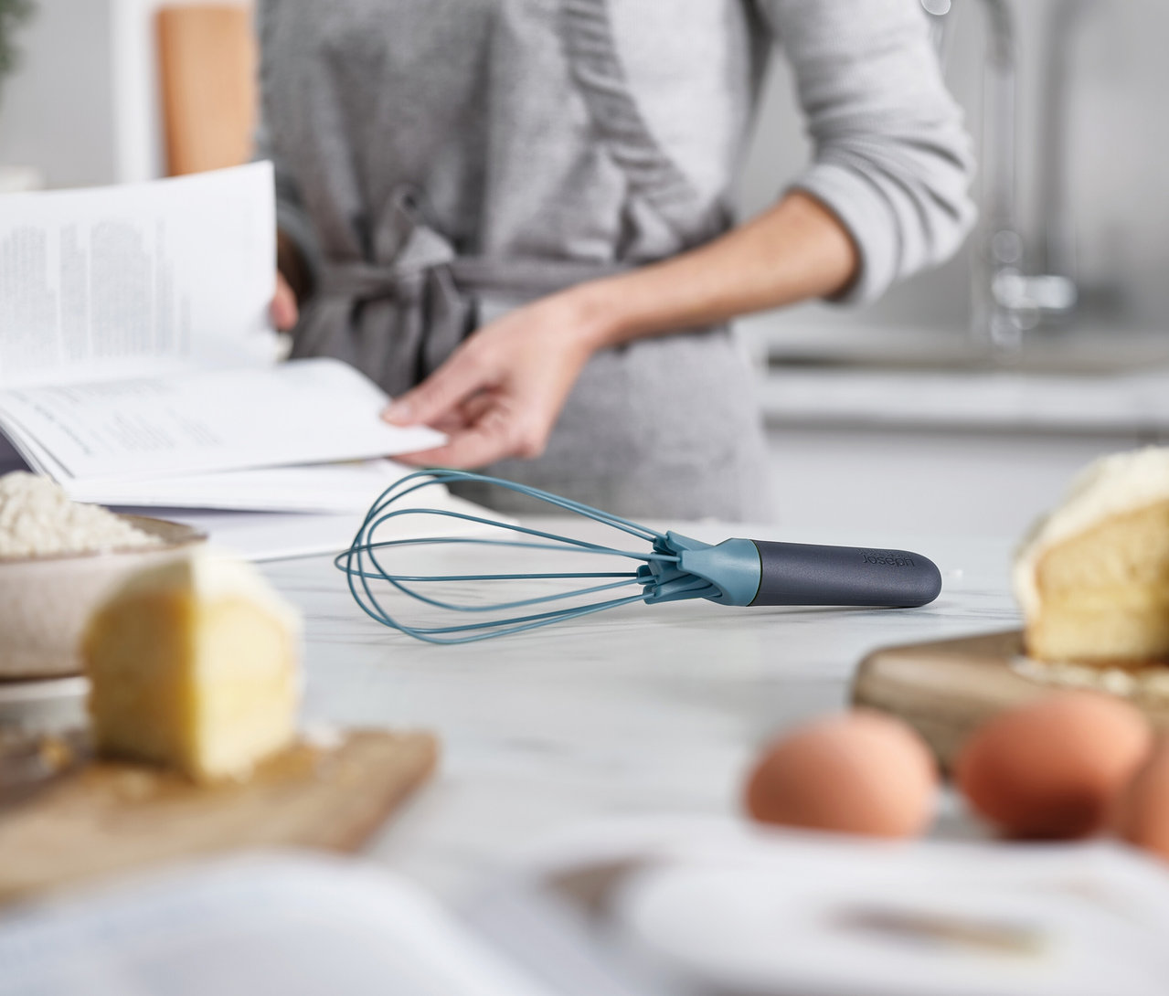 Eine Person liest ein Rezeptbuch in der Küche. Im Vordergrund liegen Eier, Butter und ein blauer Schneebesen auf einer Küchentheke.