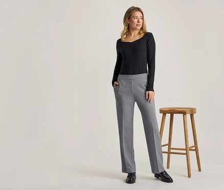 Eine blonde Frau steht in einem schwarzen Langarmshirt und einer grauen Hose, eine Hand in der Tasche, neben einem Holzhocker.