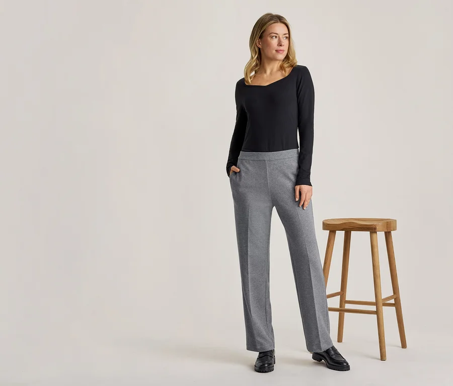 Eine blonde Frau steht in einem schwarzen Langarmshirt und einer grauen Hose, eine Hand in der Tasche, neben einem Holzhocker.