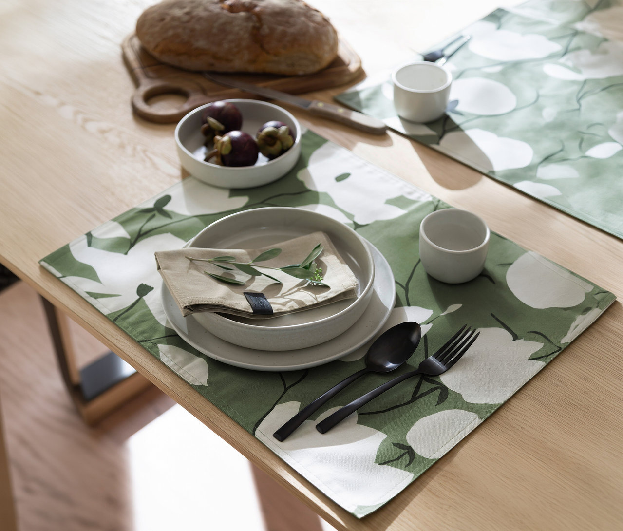 Ein Holztisch ist für eine Mahlzeit gedeckt, mit zwei khaki-floralen SCHÖNER WOHNEN-Kollektion Tischsets. Auf dem vorderen Tischset stehen ein Stapel hellgrauer Teller mit einer beigen Serviette und einem grünen Zweig, schwarzes Besteck und eine weiße Tasse. Eine Schale Mangostanen, ein Brotlaib auf einem Schneidebrett und eine weitere weiße Tasse befinden sich ebenfalls auf dem Tisch.