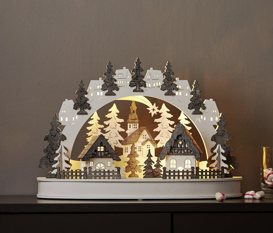 Beleuchteter Holzschwibbogen mit einer Winterlandschaft aus Häusern, einer Kirche und Bäumen. Der weiße Bogen hat dunkelgraue Bäume und Häuser mit leuchtenden Fenstern, auf einer dunklen Oberfläche.