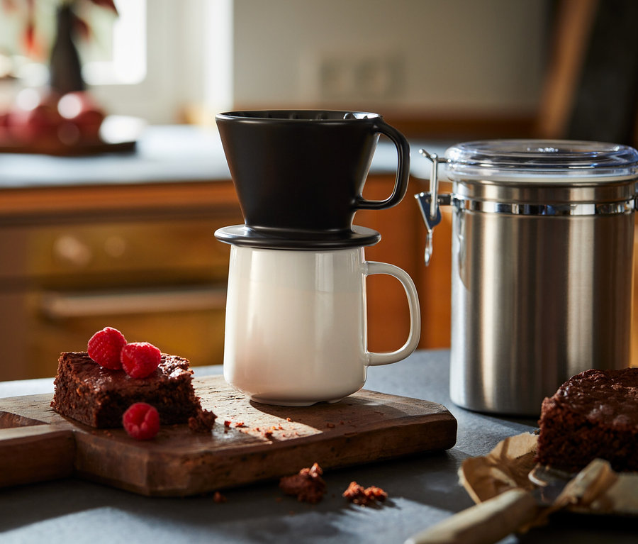 Ein schwarzer Kaffee Handfilter der Größe 101 steht auf einer weißen Tasse neben Brownies mit Himbeeren auf einem Holzbrett.