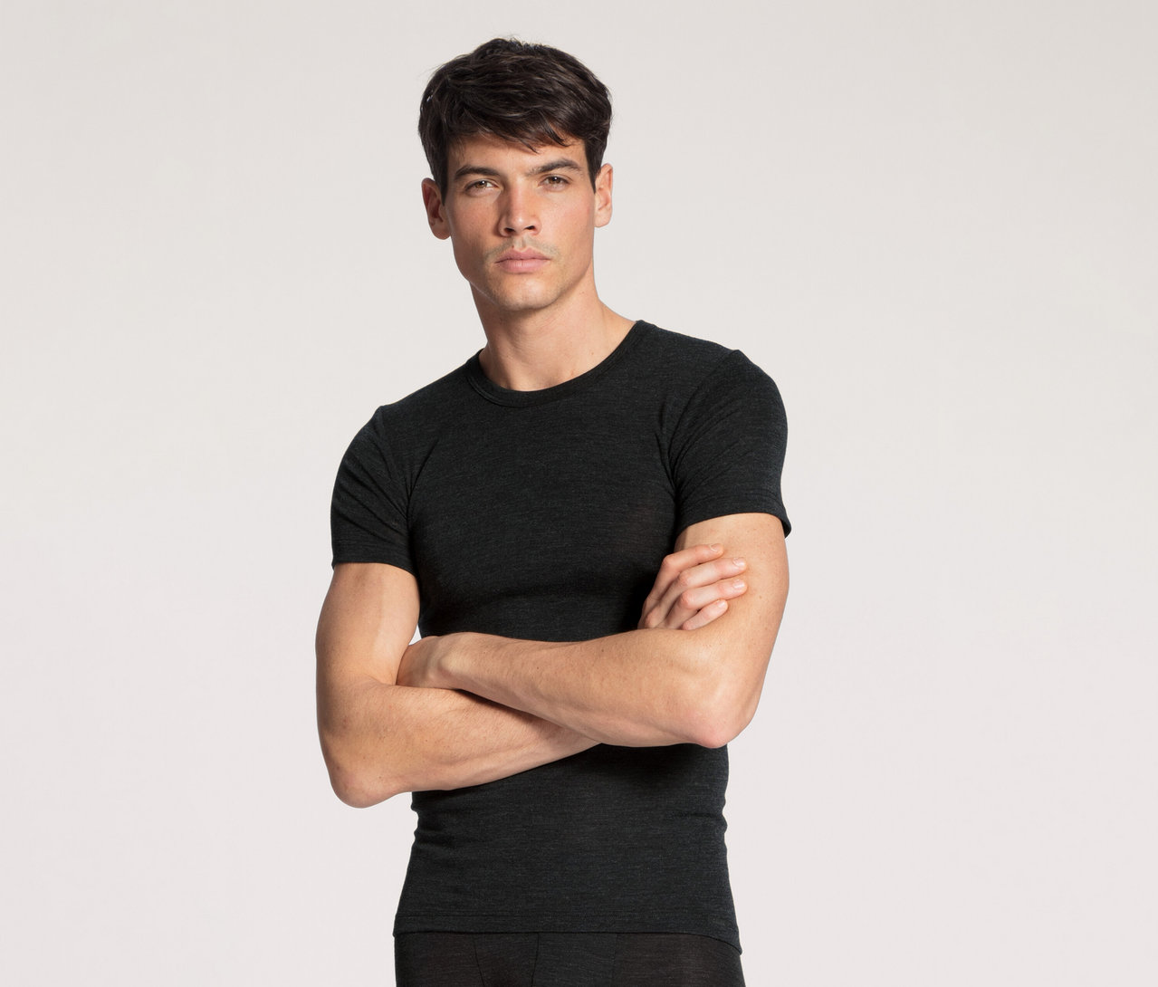 Ein Mann mit dunklem Haar und neutralem Ausdruck steht frontal da, trägt ein dunkelgraues Kurzarm-T-Shirt und dunkle Unterwäsche/Hose. Seine Arme sind vor der Brust verschränkt.
