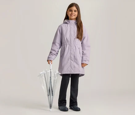 Ein lächelndes Mädchen in einem lila Mantel, dunklen Hosen und dunklen Schuhen hält einen geschlossenen, durchsichtigen Regenschirm.