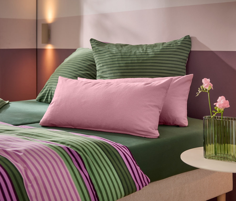 Ein Bett mit grünem Spannbetttuch, grün gestreiften Kissen, rosa Kissen und einer gestreiften Bettdecke. Ein weißer Beistelltisch trägt eine grüne gerippte Vase mit rosa Blumen. Die Wand dahinter hat horizontale rosa und lila Streifen mit zwei Wandleuchten.