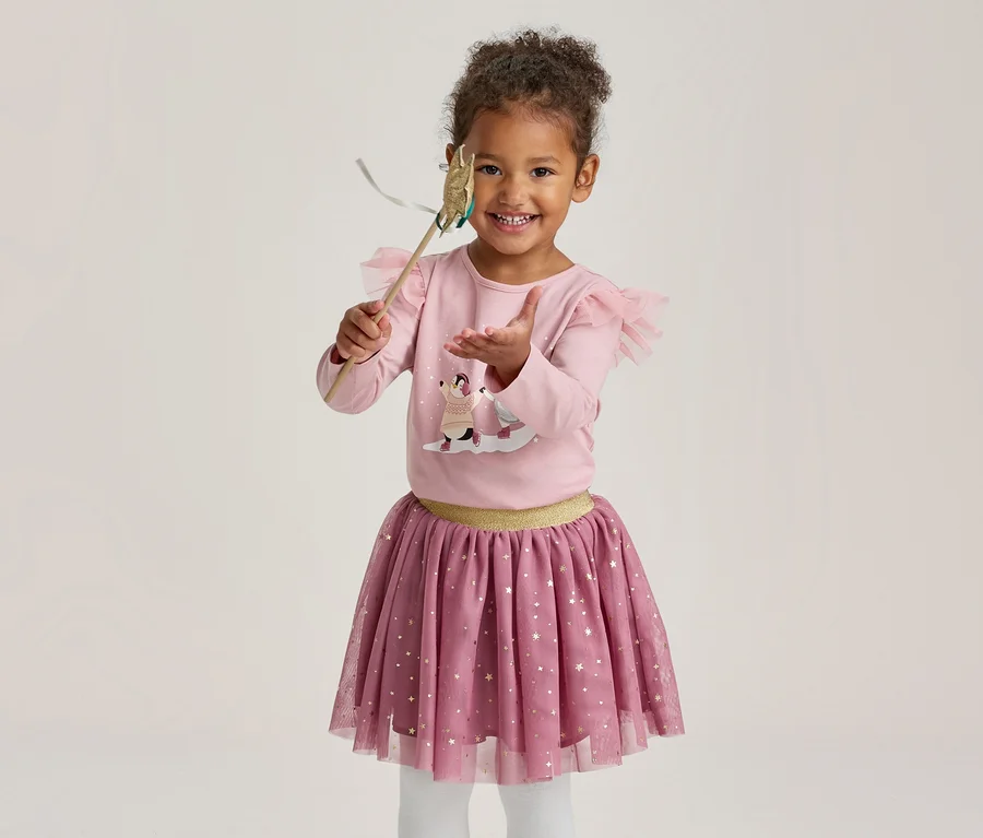 Ein lächelndes kleines Mädchen in einem rosa Langarmshirt mit Motiv und Rüschen, einem rosa Tüllrock mit goldenen Sternen und weißer Strumpfhose, das einen Spielzeugzauberstab hält.