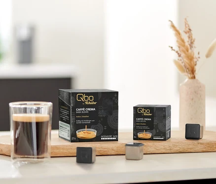 Auf einem Tisch stehen Qbo Caffè BABA BUDAN Kapseln, eine Tasse Kaffee und Kapsel-Prototypen auf einem Holzbrett.