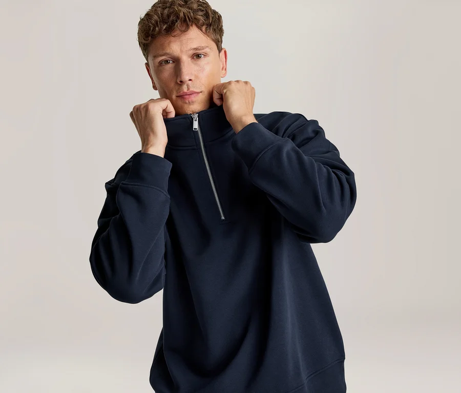 Mann in einem marineblauen Half-Zip-Sweatshirt, der den Kragen mit beiden Händen hochhält.