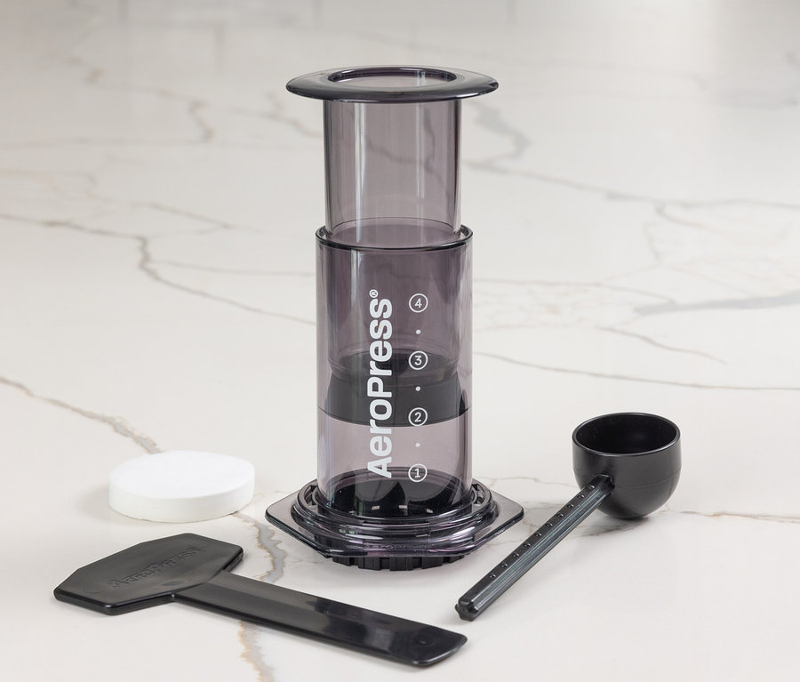 AeroPress Kaffeebereiter »Clear«, schwarz auf Marmor Hintergrund. Filter, Messlöffel und Rührstab liegen daneben.