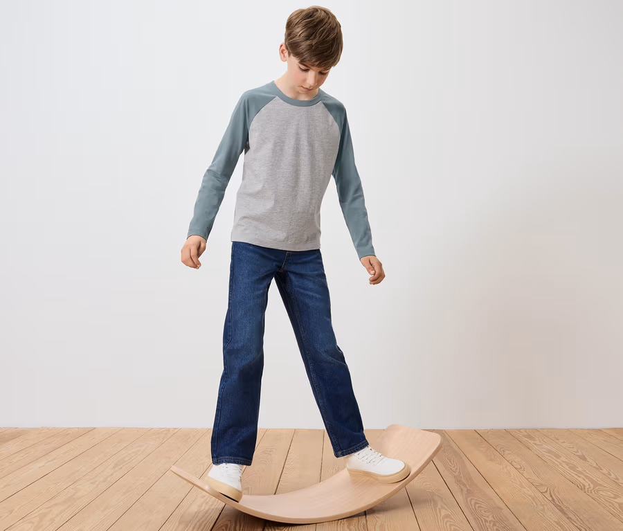 Junge steht auf einem hölzernen Balance Board, trägt ein graues T-Shirt und Jeans.