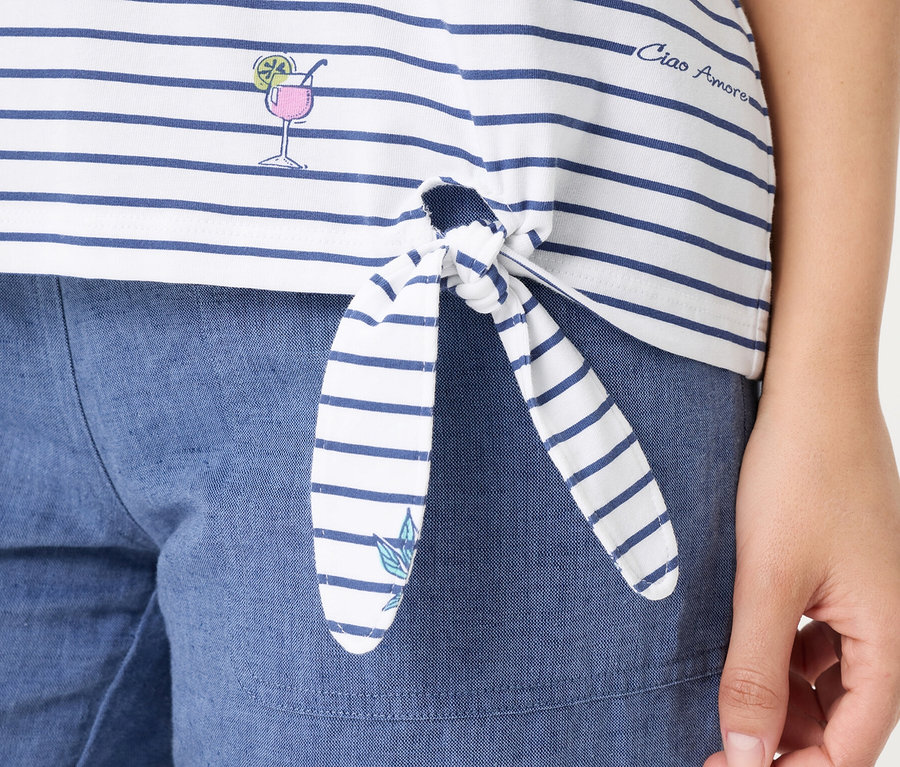 Nahaufnahme einer Person, die ein weiß-blau gestreiftes Olsen T-Shirt trägt, das vorne geknotet ist. Das T-Shirt zeigt ein gesticktes Cocktailglas, den Schriftzug 'Ciao Amore' und ein kleines Blatt am Knotenende, kombiniert mit blauen Shorts.
