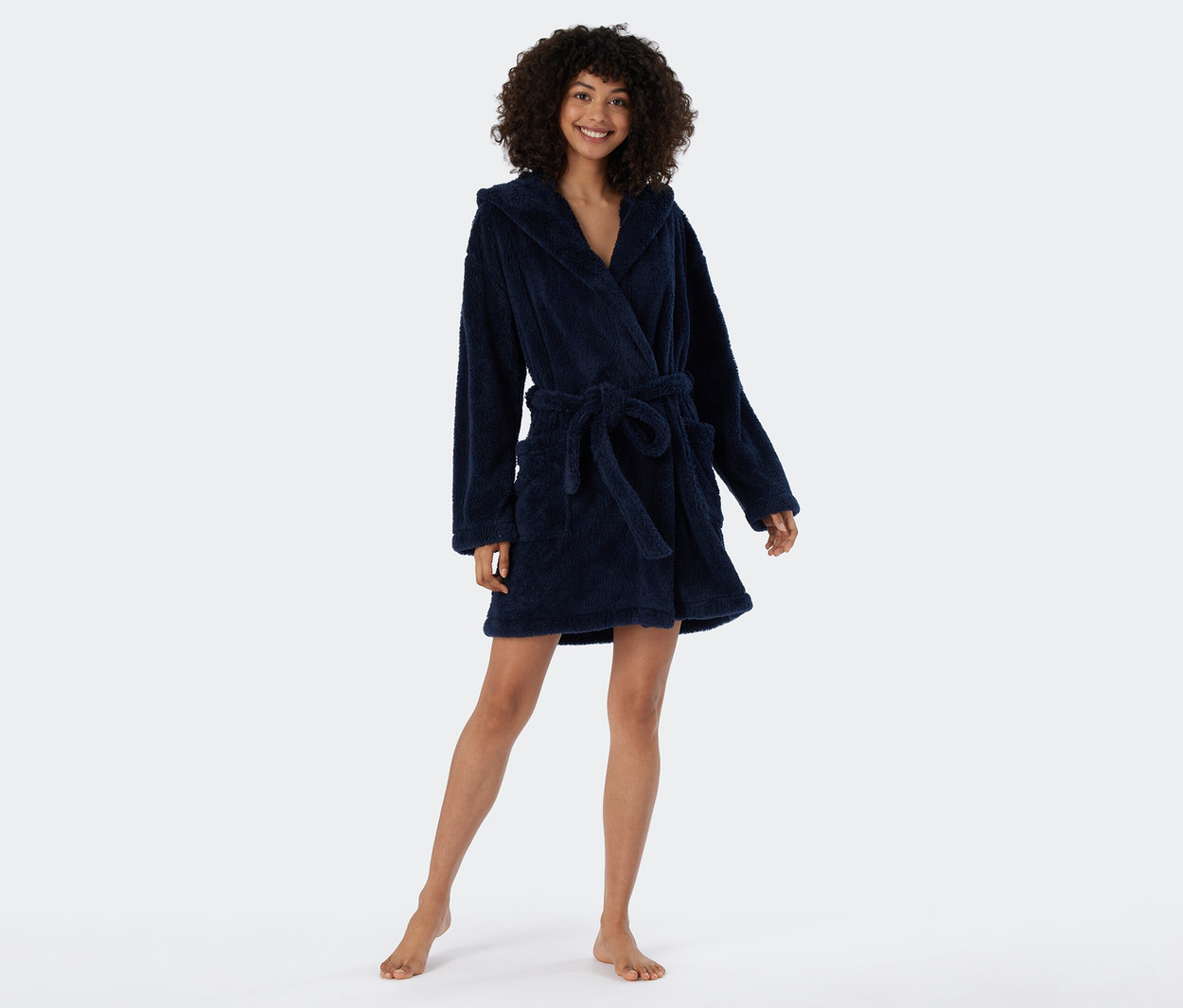 Lachende Frau mit Locken, barfuß stehend in einem marineblauen, flauschigen Bademantel mit Kapuze, in der Taille gebunden.
