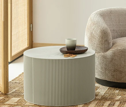 Ein runder, hellgrauer Couchtisch mit gerippter Basis steht auf einem gemusterten Teppich. Auf dem Tisch befinden sich eine dunkle Holzschale, eine zweifarbige graue Tasse und mehrere Kekse. Ein beiger Sessel und ein hölzerner Raumteiler sind teilweise sichtbar.