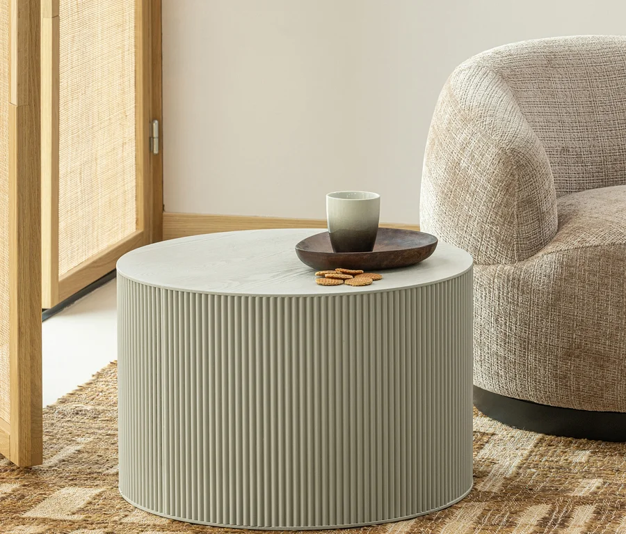 Ein runder, hellgrauer Couchtisch mit gerippter Basis steht auf einem gemusterten Teppich. Auf dem Tisch befinden sich eine dunkle Holzschale, eine zweifarbige graue Tasse und mehrere Kekse. Ein beiger Sessel und ein hölzerner Raumteiler sind teilweise sichtbar.