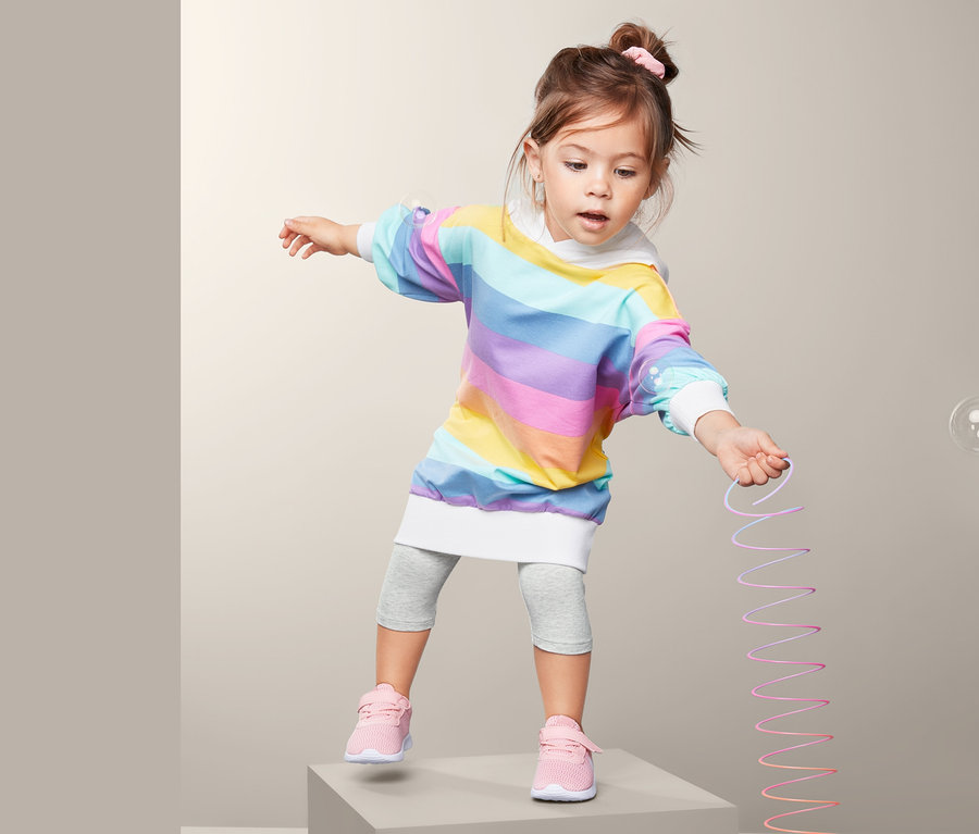 Mädchen steht auf einem grauen Würfel und hält ein Dehnspielzeug. Sie trägt ein Regenbogenkleid und 2 Leggings, 3/4-Länge.