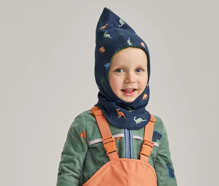 Lächelnder Junge mit blauem Dino-Balaclava, grünem Fleecepullover und orangefarbener Latzhose vor grauem Hintergrund.