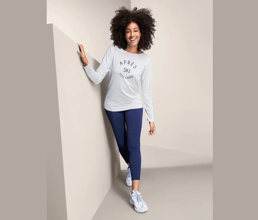 Eine Frau mit lockigem Haar lächelt und posiert in einem weißen Funktionsshirt mit langen Ärmeln und dunkelblauen Leggings.
