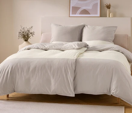 Ein Bett mit taupe-offweißer Flanell-Bettwäsche. Der Bettbezug hat oben und unten taupefarbene Bereiche mit einem offweißen Mittelstreifen, zwei taupefarbene Kissen liegen an einem hellen Kopfteil.